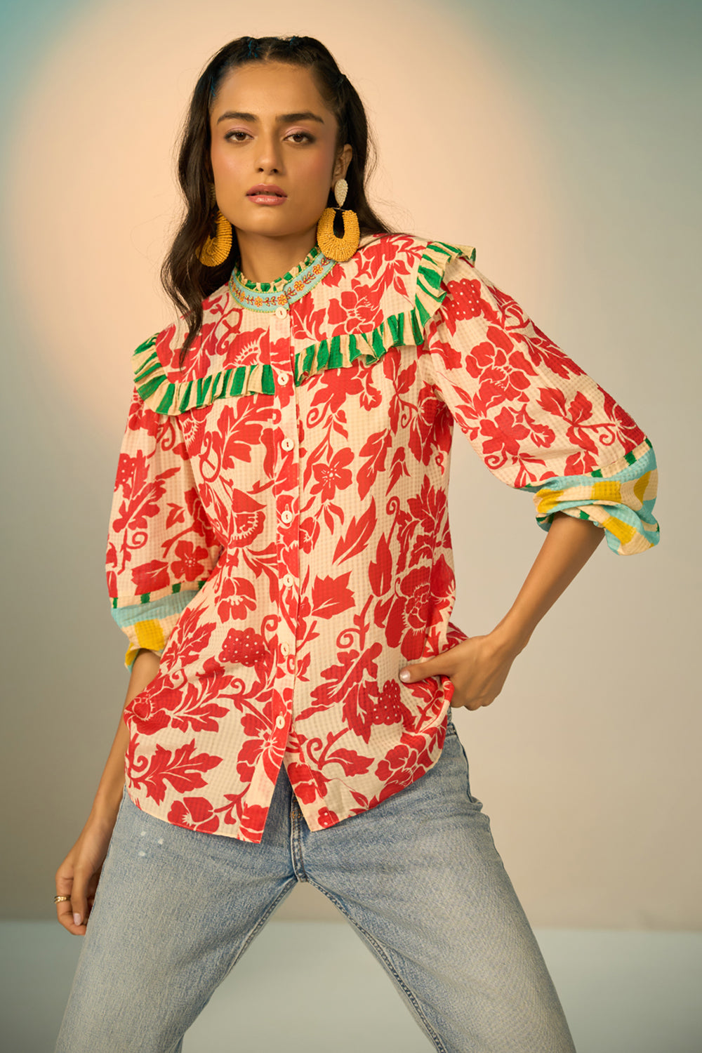 A woman modeling a vibrant red and cream floral print blouse with green and yellow accents, paired with light-wash jeans and gold hoop earrings, set against a soft beige studio background.