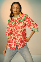 A woman modeling a vibrant red and cream floral print blouse with green and yellow accents, paired with light-wash jeans and gold hoop earrings, set against a soft beige studio background.