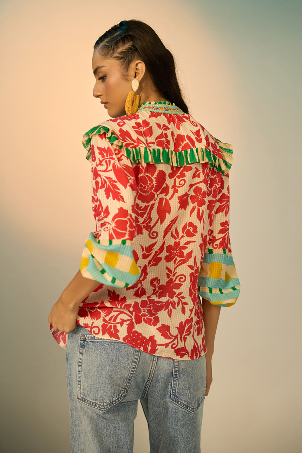 A woman modeling a vibrant red and cream floral print blouse with green and yellow accents, paired with light-wash jeans and gold hoop earrings, set against a soft beige studio background.