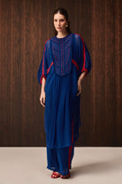 A woman models a royal blue tunic and trouser set with red and navy embroidered details on the chest and red piping along the sleeves and side seams, set against a dark wood-paneled background.