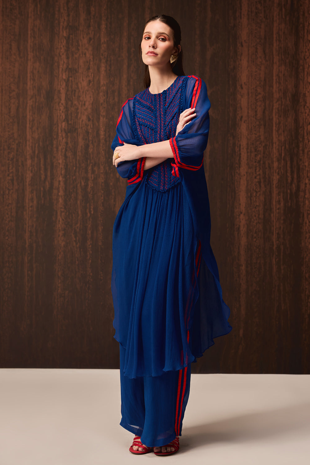 A woman models a royal blue traditional outfit with red embroidered details and red side stripes, standing against a dark wood-paneled background.