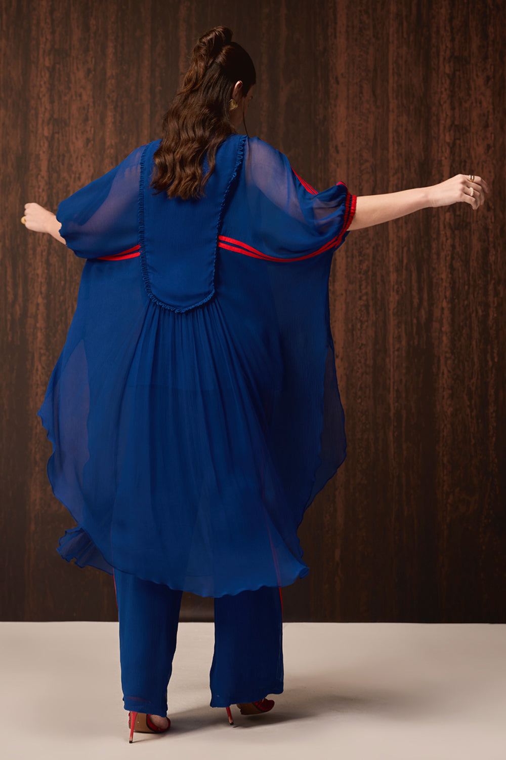 A woman viewed from behind wearing a flowing royal blue sheer tunic with red trim and matching blue pants, posed against a dark wood-paneled background.