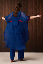 A woman viewed from behind wearing a flowing royal blue sheer tunic with red trim and matching blue pants, posed against a dark wood-paneled background.