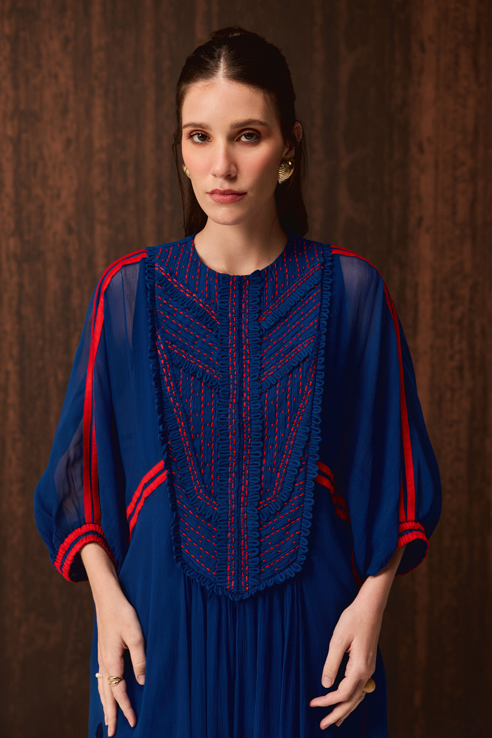 A woman wearing a royal blue dress with red embroidered detailing on the chest and red stripes on the sleeves, posed against a dark brown textured background.