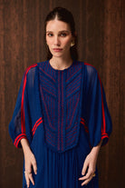 A woman wearing a royal blue dress with red embroidered detailing on the chest and red stripes on the sleeves, posed against a dark brown textured background.