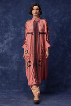 A woman models a pink long kurta with green floral embroidery and dark red and blue vertical stripe details, paired with patterned trousers, against a textured blue studio backdrop.