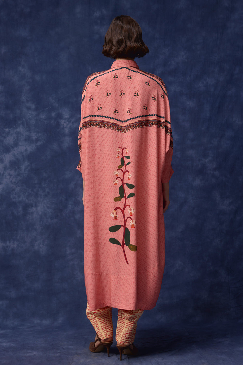 A model viewed from behind wearing a dusty pink shawl with black geometric embroidery on the shoulders and a vertical green floral vine design on the back, set against a textured blue studio backdrop.