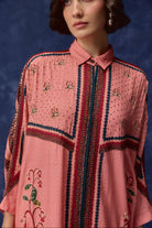 A close-up of a woman wearing a pink collared shirt with intricate red, navy, and green floral embroidery and geometric border patterns, set against a dark blue textured background.