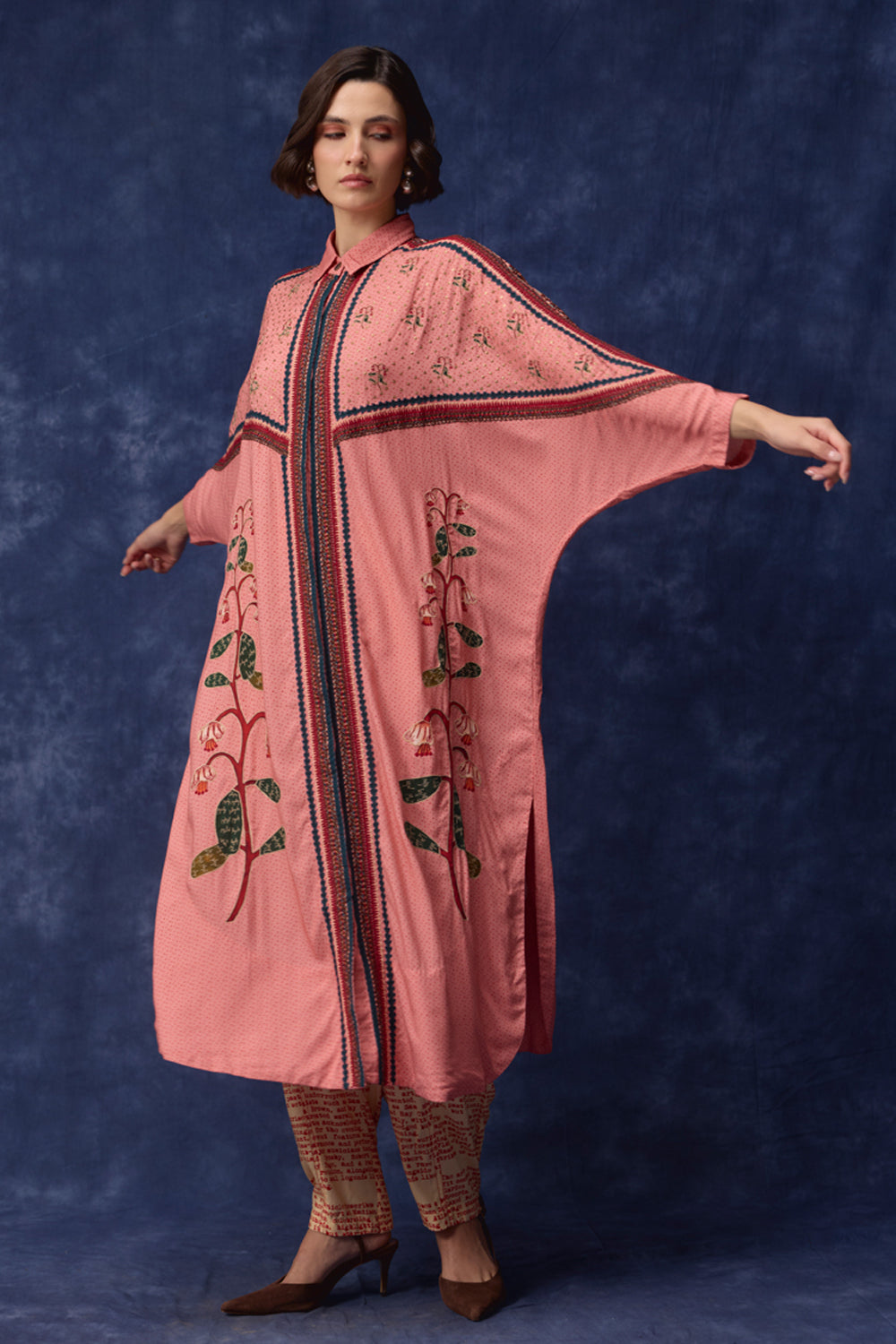 A woman modeling a pink embroidered kaftan dress with wide sleeves, standing against a textured dark blue studio backdrop.