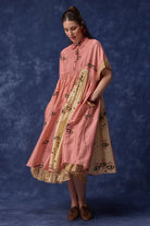 A woman modeling a pink and beige tiered dress with floral embroidery and patterned borders, standing against a textured blue studio backdrop.