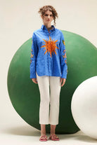 Arin cobalt blue cotton shirt with relaxed fit, long sleeves and embroidered orange sun motif styled with white trousers