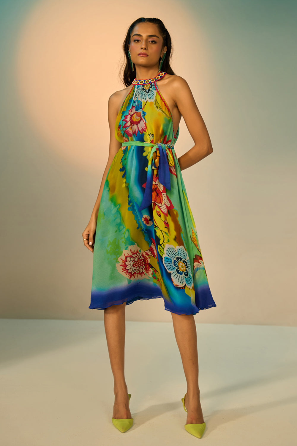 A woman modeling a sleeveless halter-neck midi dress with a vibrant multicolor floral and abstract print in shades of blue, green, yellow, and pink, styled with lime green pointed-toe heels against a studio backdrop.