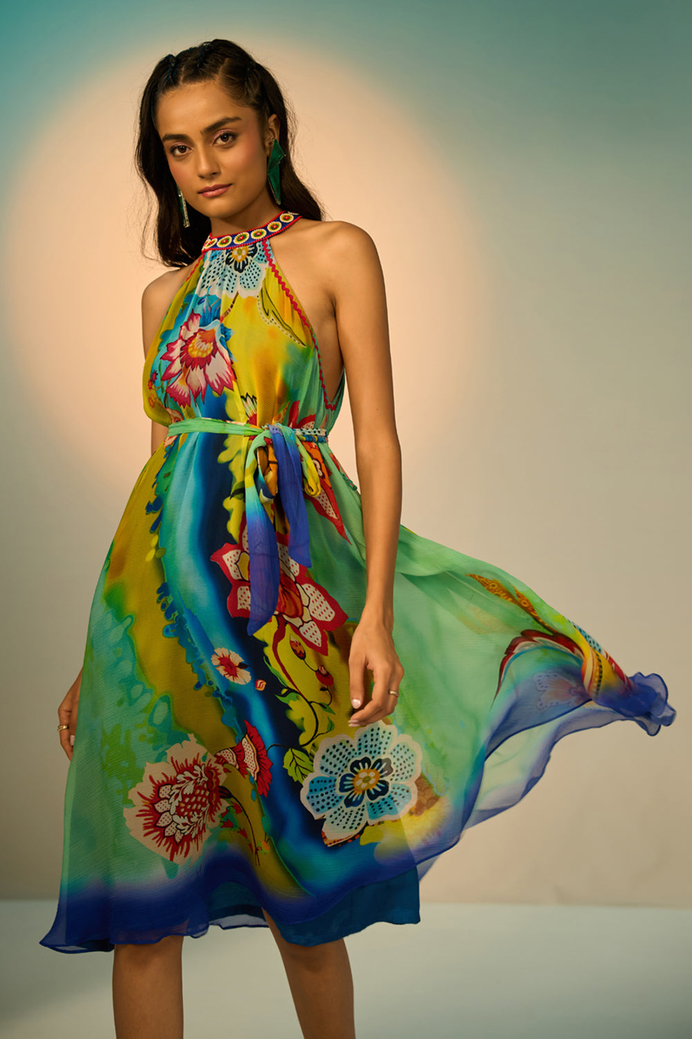 A woman modeling a sleeveless halter-neck midi dress with a vibrant multicolor floral and abstract print in shades of blue, green, yellow, and pink, styled with lime green pointed-toe heels against a studio backdrop.