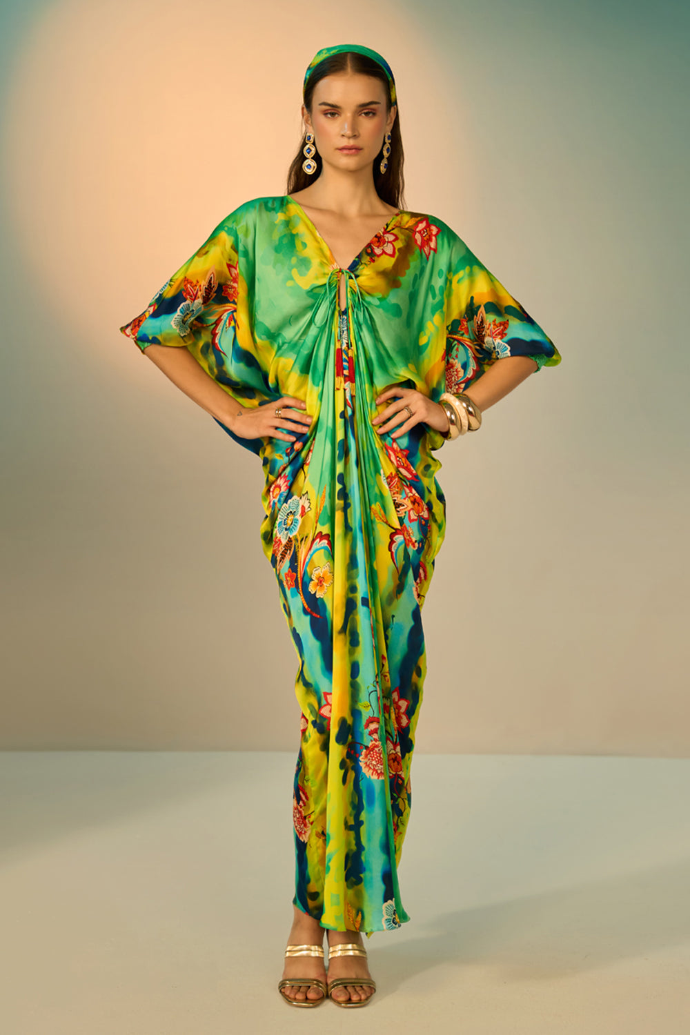A woman modeling a vibrant, multicolored kaftan dress with a tie-dye pattern in multicolor, styled with a matching headband and gold accessories against a neutral studio backdrop.