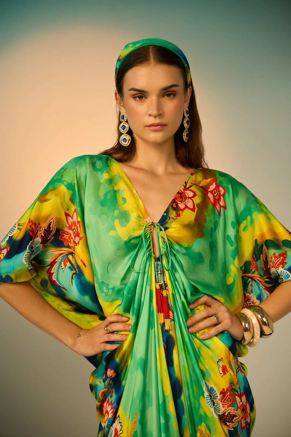 A close-up loof of woman modeling a vibrant, multicolored kaftan dress with a tie-dye pattern in multicolor, styled with a matching headband and gold accessories against a neutral studio backdrop.