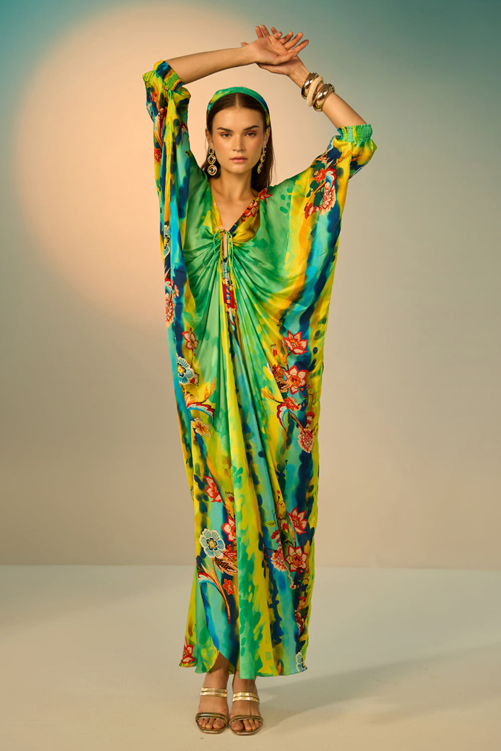 A woman modeling a vibrant, multicolored kaftan dress with a tie-dye pattern in multicolor, styled with a matching headband and gold accessories against a neutral studio backdrop.