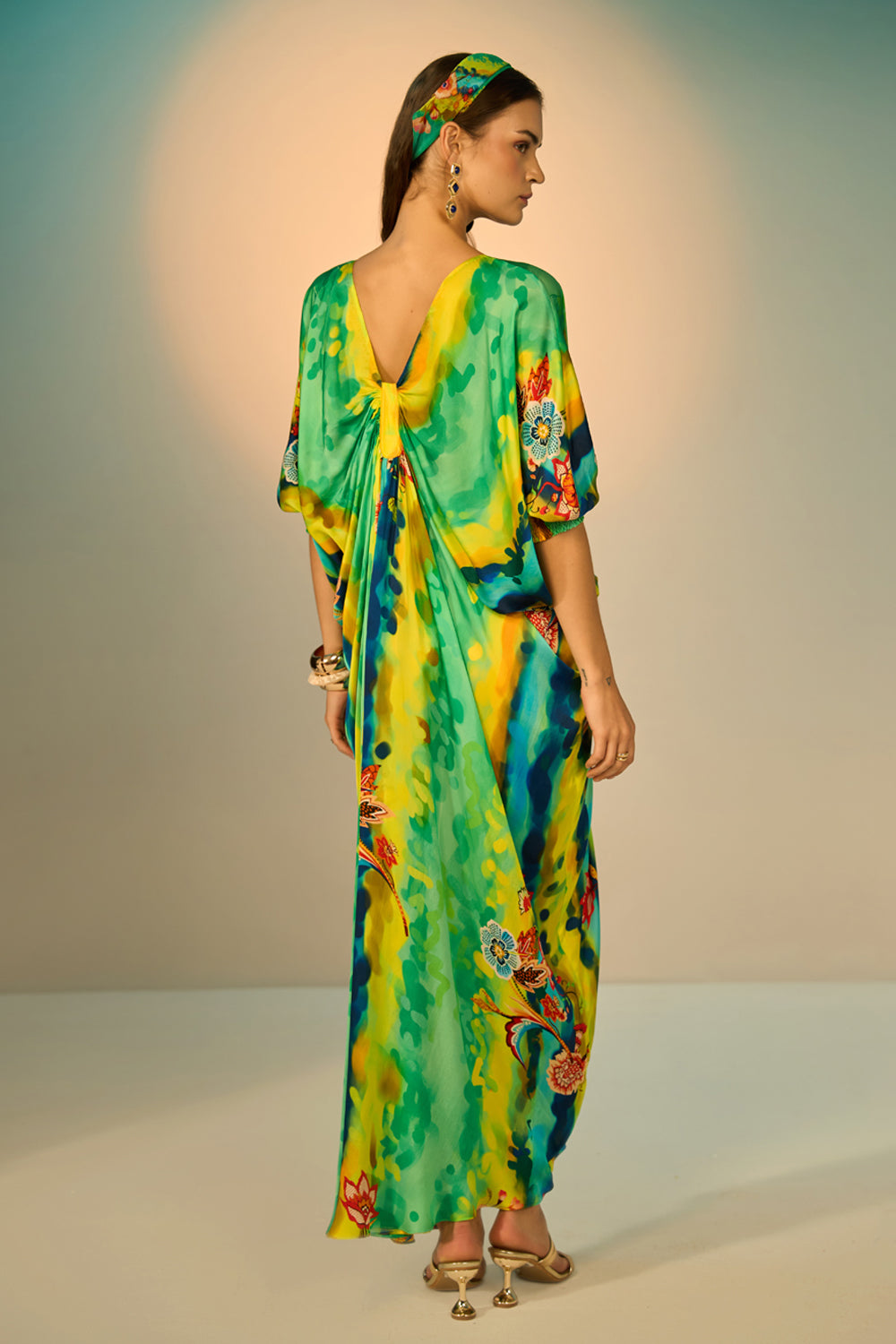 Back view of a woman modeling a vibrant, multicolored kaftan dress with a tie-dye pattern in multicolor, styled with a matching headband and gold accessories against a neutral studio backdrop.