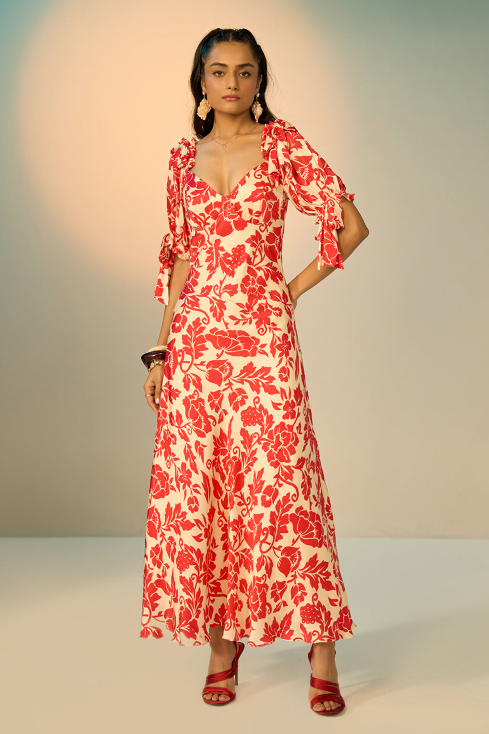 A woman modeling a red and cream floral print maxi dress with puff sleeves and a sweetheart neckline, standing against a neutral studio backdrop.
