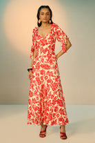 A woman modeling a red and cream floral print maxi dress with puff sleeves and a sweetheart neckline, standing against a neutral studio backdrop.