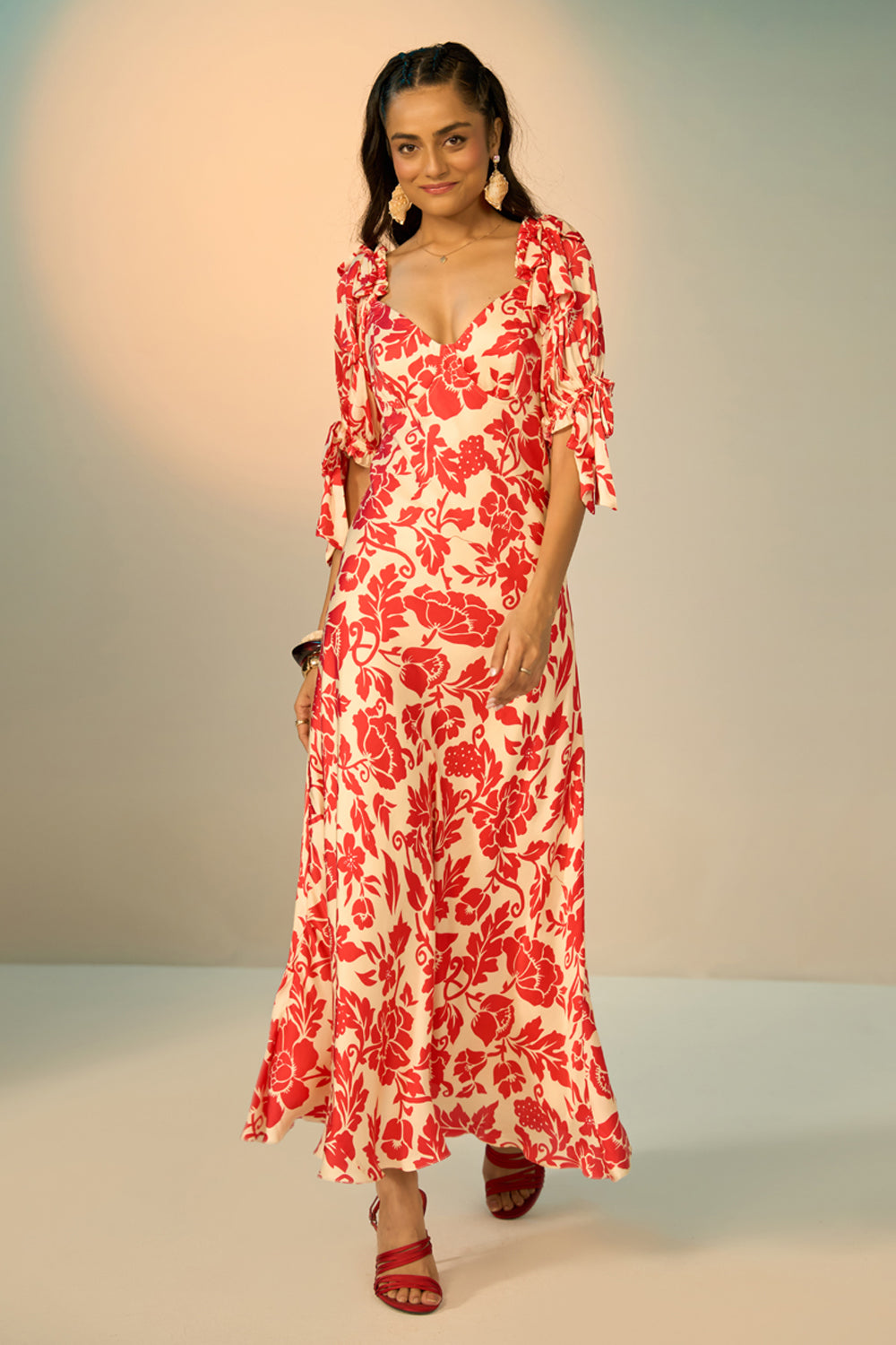 A woman modeling a red and cream floral print maxi dress with puff sleeves and a sweetheart neckline, standing against a neutral studio backdrop.