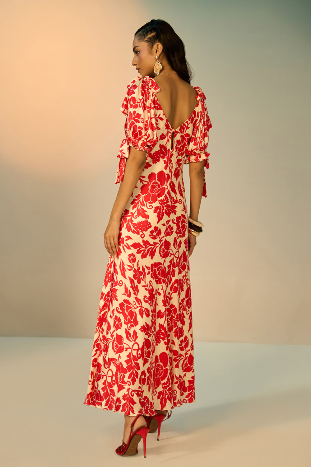 A woman modeling a red and cream floral print maxi dress with puff sleeves and a sweetheart neckline, standing against a neutral studio backdrop.